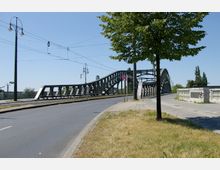 Eine Stahlbrücke mit geschwungenem, offenen Geländer überquert eine Straße und ein benachbartes Gleis. Im Vordergrund befinden sich ein Gehweg, ein einzelner Baum und Straßenlaternen, während der Himmel klar und blau ist.