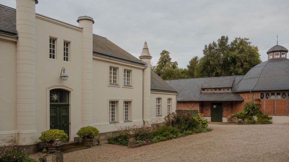 Innenhof eines Gutshauses mit hellem, zweigeschossigem Hauptgebäude und dunkler Eingangstür zwischen runden Pfeilervorsprüngen. Rechts stehen rote Backsteingebäude mit Schieferdach und einem runden Kuppeldach; davor ein Kiesweg und bepflanzte Beete.