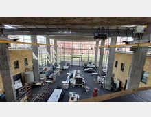 Großes Industriegebäude mit einer hohen, gläsernen Front, die Blick auf einen grünen Außenbereich und ein rotes Backsteingebäude bietet. Im Innenraum befinden sich technische Geräte, Testaufbauten und Werkbänke, umgeben von Betonpfeilern und gelben Kranträgern an der Decke.