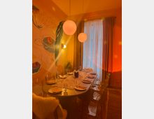 Warm-lit private dining room with a long table set for multiple guests, clear acrylic chairs, and two round pendant lights overhead. A large wall mural decorates one side, and tall curtains cover a floor-to-ceiling window at the end of the room.