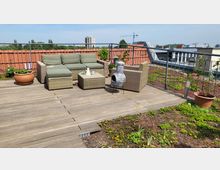 Holzterrasse mit Sitzmöbeln aus geflochtenem Material und grünen Polstern, einem kleinen Tisch und einer dekorativen Feuerschale. Im Vordergrund eine bepflanzte Dachfläche, im Hintergrund Dachziegel, Geländer und Blick auf umliegende Gebäude sowie einen Kran in der Ferne.