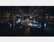 Industriehalle mit einer Bühne in der Mitte, umgeben von Metallträgern und Beleuchtungselementen. Auf der Bühne sind mehrere Darsteller und kreisförmige Konstruktionen, während im Vordergrund leere Sitzreihen für Zuschauer zu sehen sind.