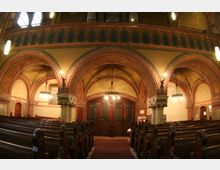 Innenraum einer Kirche im neogotischen Stil mit roten Backsteinbögen und verzierten Säulen. Sichtbare Elemente umfassen Holzbänke, kunstvolle Leuchter und eine Holztür mit Glasdetails, umgeben von detaillierten Wand- und Deckenmalereien.