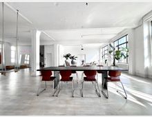 Weitläufiger, heller Arbeits- oder Wohnraum mit hohen Decken, großen Fenstern und weißen Wänden. Im Vordergrund steht ein langer Tisch mit roten Stühlen, im Hintergrund sind Pflanzen, eine Couch und ein Spiegel an der Wand sichtbar.