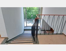 Treppenhaus in einem Gebäude mit schlichtem Design, bestehend aus einer Treppe mit schwarzem Metallgeländer und roten Handläufen. Am Ende des Treppenabsatzes befinden sich große Fenster, die Tageslicht hereinlassen und den Blick auf grüne Bäume und einen gepflasterten Außenbereich freigeben.