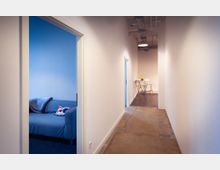 Ein schmaler, weiß gestrichener Flur mit Betonboden führt zu zwei geöffneten Türen. Links befindet sich ein Raum mit einem blauen Sofa und einem bunten Kissen, rechts ein kleiner Essbereich mit Klapptisch und -stühlen auf dunklem Holzboden.