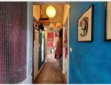 Schmaler Flur in einer Wohnung mit blauen Wänden, Holzboden und hängender Wanddekoration, darunter gerahmte Bilder. An den Wänden hängen Kleidung und Hüte, im Hintergrund ein Wohnraum mit roten Wänden und Möbeln sichtbar.