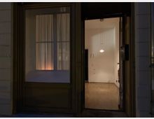 Eine geöffnete Eingangstür eines Gebäudes bei Nacht, die einen beleuchteten Innenraum mit Holzfußboden, weißen Wänden und einer hängenden Deckenlampe zeigt. Links neben der Tür befindet sich ein großes Fenster mit geschlossenen Vorhängen.