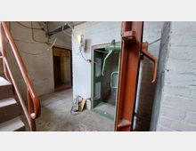 Industrieartiger Kellerbereich mit weißen Backsteinwänden, einem offenen Treppenaufgang links und einem grünen, schweren Stahltürrahmen rechts. An der Wand hängen freiliegende Kabel und Rohre, und eine Glühbirne beleuchtet schwach den Raum.