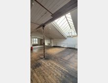 Großer, leerer Dachgeschossraum mit breitem Oberlicht aus Glas im schrägen Dach, weißen Wänden und sichtbaren Holzbalken. Alter Holzdielenboden, mehrere Heizkörper entlang der Wände und eine einzelne Stützsäule in der Raummitte.