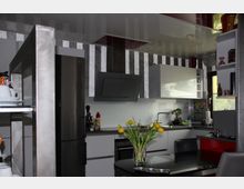 Moderne Küche mit weißen Schränken, grauer Arbeitsplatte und einer Edelstahl-Dunstabzugshaube. Im Vordergrund steht ein Esstisch mit einer Glasvase, die mit gelben Tulpen dekoriert ist, und im Hintergrund ist ein großes Fenster sowie ein Kühlschrank zu sehen.