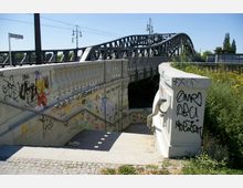 Treppenabgang zu einer Unterführung einer Brücke mit Geländern aus Beton, übersät mit Graffiti. Im Hintergrund ist eine Stahlbrücke mit dekorativen Lampen und umliegender Vegetation zu sehen.