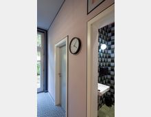 Ein Innenraum mit einer rosa Wand, an der eine analoge Wanduhr hängt, einer Tür zu einem Raum mit schwarz-weißem Musterboden, und einer weiteren offenen Tür zu einem Badezimmer mit grün-schwarz gemusterten Fliesen, einem Waschbecken und einem Seifenspender. Links ist eine verglaste Tür mit Blick nach draußen sichtbar.