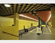 U-Bahn-Station mit gelb gefliesten Wänden und einem Schachbrettmuster an der Decke. Die Station hat zwei Bahnsteige, eine gelbe U-Bahn hält rechts; Hinweisschilder und Menschen sind im Bereich der Bahnsteige sichtbar.