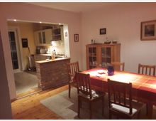 Esszimmer mit einem Tisch, der mit einer rot-orange karierten Tischdecke bedeckt ist und von Holzstühlen umgeben ist. Im Hintergrund ist eine offene Küche mit gelben Wänden, weißen Hängeschränken und einer Theke mit Backsteinverkleidung zu sehen.