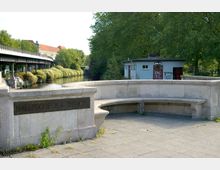 Steinerne Sitzbank mit der Inschrift „Hallesche-Tor-Brücke“ im Vordergrund, dahinter ein kleiner Fluss mit Ufervegetation und ein Gebäudeschuppen mit Graffiti. Auf der linken Seite verläuft eine erhöhte Eisenbahn- oder U-Bahn-Brücke parallel zum Fluss.