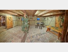 Innenraum eines rustikalen Fachwerkgebäudes mit Ziegelboden und Holzbalkendecke. Es befinden sich Holztische, Stühle und Dekorationen wie getrocknete Pflanzen und landwirtschaftliche Werkzeuge an den Wänden.