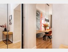 Blick durch einen Flur in einen modernen Essbereich mit Holzboden, großem Holztisch und braunen Stühlen. Über dem Tisch hängen mehrere schwarze Pendelleuchten, an der Wand ist ein großformatiges Wandbild, und seitlich steht eine schmale Konsole mit Vase.