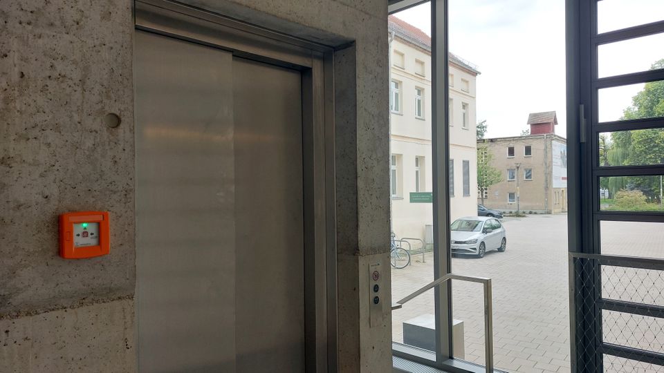 Ein moderner Innenraum mit einem Aufzug aus Edelstahl in einer Betonwand; daneben ein orangefarbener Notrufschalter. Durch große Fensterfronten ist ein Blick auf einen gepflasterten Hof mit parkenden Autos und umliegenden Gebäuden sichtbar.