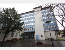 Moderner Büro- oder Verwaltungsbau mit weißer Fassade und großen rechteckigen Fenstern. Der Eingangsbereich ist mit Glas gestaltet, davor befindet sich ein gepflasterter Hof mit Bäumen, Sträuchern und einer Straßenlaterne.