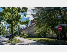Straßenszene in einem Wohngebiet mit mehrstöckigen Gebäuden mit roten Ziegeldächern auf einer Seite und einer von Bäumen gesäumten Straße. Im Vordergrund ein Gehweg, ein Straßenschild mit Stoppschild sowie parkende Autos auf der linken Straßenseite.