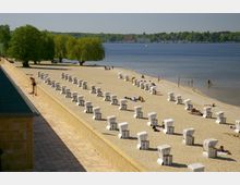 Strand mit mehreren Reihen von weißen Strandkörben auf hellem Sand, flankiert von einem gepflasterten Weg und Bäumen. Im Hintergrund ein See mit ruhigem Wasser und einer bewaldeten Uferlinie sowie vereinzelte Badegäste am Strand.