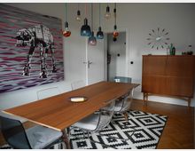 Esszimmer mit modernem Design: Ein langer Holztisch wird von transparenten Stühlen umgeben, darüber hängen dekorative Hängeleuchten in verschiedenen Farben und Formen. An der Wand befindet sich ein großes Kunstwerk mit einem stilisierten AT-AT-Motiv, daneben steht ein Vintage-Holzschrank, und der Boden ist mit einem Teppich im schwarz-weißen geometrischen Muster bedeckt.