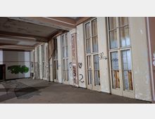 Verlassener Innenraum eines Gebäudes mit einer langen Reihe hoher, mehrfach verglaster Türen und Fenster, teils beschädigt und mit abblätternder Farbe. An Wänden und Türen sind Graffiti zu sehen; der Boden ist stellenweise verschmutzt und mit Trümmern bedeckt.
