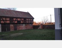 Fachwerkscheune aus rotem Ziegelstein mit schwarzem Holzgebälk und abgenutzten Türen, umgeben von einem grasbewachsenen Hof. Im Hintergrund stehen ein Holztor, ein Baum und eine teils verputzte Mauer, dahinter sind weitere Bäume und eine ländliche Szenerie sichtbar.