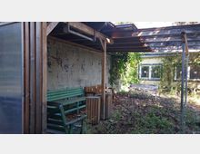 Überdachter Sitzbereich mit Holzbänken und Abfalleimern auf einem Boden aus Laub und Pflanzenresten. Im Hintergrund ist eine mit Graffiti bemalte Wand zu sehen sowie ein Gebäude mit gelber Fassade und Fenstern, das von dichter Vegetation umgeben ist.