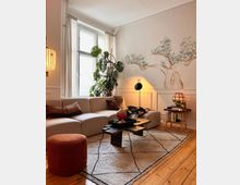 Wohnzimmer mit einer hellen Sofalandschaft vor einem großen Fenster mit weißen Vorhängen. An der Wand ist eine Malerei eines Baumes zu sehen, flankiert von einer großen Zimmerpflanze. Ein moderner dunkler Couchtisch steht auf einem gemusterten Teppich, daneben ein roter Pouf und eine Stehlampe. Der Holzfußboden und warme Beleuchtung sorgen für eine gemütliche Atmosphäre.