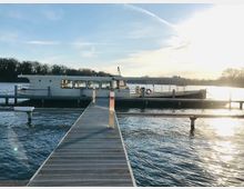 Ein Boot liegt an einem Steg vor Anker auf einem See, umgeben von ruhigem Wasser und Bäumen im Hintergrund. Die Szene ist in warmes Licht der tiefstehenden Sonne getaucht.