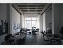 Großer, heller Büroraum mit hohen Decken und sichtbaren Betonträgern. Der Raum ist mit mehreren Schreibtischen, Stühlen und Büromaterialien ausgestattet; eine Fensterfront am Ende des Raumes lässt viel Tageslicht herein.