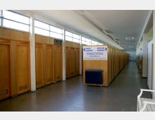 Langer, schlichter Umkleideraum mit einer Reihe hölzerner Kabinentüren, nummeriert von 1 bis 7. Oben befinden sich durchgehende Fenster, die Tageslicht einlassen, und in der Mitte steht ein Schild mit Hinweisen zu „Dauer“ und „Wechselkabinen“.