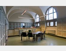 Ein historischer Raum mit gewölbter Decke, großen Bogenfenstern und verzierten Wandpaneelen mit floralen Mustern. Im Vordergrund stehen ein Tisch und mehrere Stühle, während im Hintergrund eine erhöhte Theke zu sehen ist.