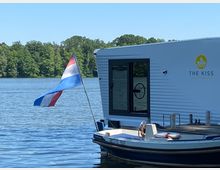 Ein weißes Hausboot mit der Aufschrift „The Kiss Klostersee“ liegt am Wasser, daneben ein kleines Motorboot. Im Hintergrund ist ein See mit bewaldetem Ufer und blauem Himmel zu sehen, rechts weht eine niederländische Flagge.