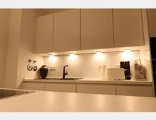 Moderne Küchenzeile mit weißen Oberschränken, beleuchteter Marmoroptik-Rückwand und weißer Arbeitsfläche. Zu den sichtbaren Elementen gehören eine schwarze Armatur, eine schwarze Vase, ein Wasserkocher, ein Toaster, Bücher und eine Kaffeemaschine.