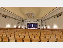 Großer Veranstaltungssaal mit Reihen von hellen Holzstühlen, einer Bühne mit violettem Vorhang, einem Rednerpult und einem Klavier. Die Wände sind weiß, mit eingelassenen Lichtpaneelen, und an der Decke befindet sich eine ovale Verzierung.