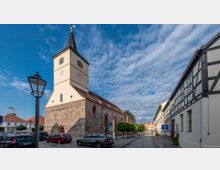 Straßenansicht mit einer Kirche aus hellem Putz und einem rechteckigen Turm mit Spitzdach und Ziffernblättern an den Uhrwerken. Rechts ein Fachwerkhaus, links parkende Autos, Kopfsteinpflaster auf der Straße und ein klarer Himmel im Hintergrund.