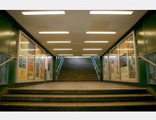 Unterführung mit Treppenaufgang, flankiert von Wänden mit grünem Metall und Schaukästen, die mit Plakaten und Anzeigen gefüllt sind. Der Bereich ist von rechteckigen Leuchtstofflampen an der Decke beleuchtet.