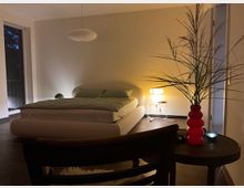 Modernes Schlafzimmer mit niedrigem, gepolstertem Bett und grüner Bettwäsche, Hängelampe sowie Stehlampe neben dem Bett. Große Fensterflächen, dunkler Boden und ein Beistelltisch mit roter Vase und hohen Zweigen im Vordergrund.