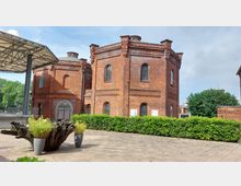 Historisches Backsteingebäude mit Rundbogenfenstern und verzierten Fassadenelementen, umgeben von einer Hecke und einem gepflasterten Platz; links ein modernes, überdachtes Metallgerüst und vorne Blumenkübel.