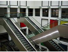 Innenansicht eines modernen Gebäudes mit mehreren Rolltreppen, die sich kreuzen. Die Wände sind weiß, mit geometrischen Fensterausschnitten und bunten Wandpaneelen im Hintergrund.