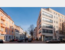 Straßenszene in einer städtischen Wohngegend mit mehrstöckigen Gebäuden auf beiden Seiten, darunter links ein orangefarbenes Gebäude mit Balkonen und rechts ein modernes Gebäude mit großen Fenstern. Am Straßenrand stehen geparkte Autos, während eine Person mit dem Fahrrad fährt.