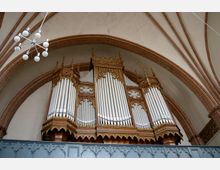 Große, historische Orgel mit reich verziertem, goldfarbenem Holzgehäuse und zahlreichen silbernen Pfeifen, eingebaut in eine gotische Kirchenarchitektur mit gewölbten Ziegelbögen. Im Vordergrund ist eine dekorativ gestaltete Holzbrüstung zu sehen.