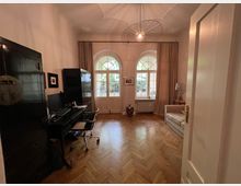 Ein heller Raum mit Holzboden, großen doppelten Bogenfenstern und hohen Decken. Links steht ein Schreibtisch mit Computer und schwarzem Schrank, rechts ein Sofa; im Hintergrund hängen Vorhänge an den Fenstern, die den Blick auf eine grüne Außenanlage freigeben.