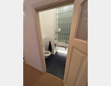 Ansicht eines kleinen Badezimmers mit geöffneter Tür, ausgestattet mit einer Toilette, einem Waschbecken und Glasbausteinen an der Wand. Der Boden besteht aus kleinen, dunklen Fliesen, und ein schwarzes Handtuch hängt an der Wand.