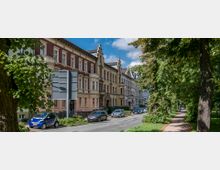 Straßenansicht mit historischer Architektur: Mehrstöckige, verzierte Altbauten in hellen Farben säumen die linke Straßenseite, während rechts ein von großen Bäumen gesäumter Parkweg zu sehen ist. Autos parken am Straßenrand, und ein Verkehrsschild steht im Vordergrund.