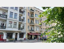 Mehrstöckige Wohnhäuser mit historischen Fassaden in einer urbanen Straßenszene, links ein restauriertes Gebäude mit weißen Balkonen und roten Fensterrahmen, rechts ein gelbliches Gebäude mit älterem Putz. Im Erdgeschoss befinden sich Geschäfte, einschließlich eines Restaurants mit Außensitzplätzen. Im Vordergrund Straßenbäume und parkende Fahrräder.