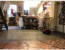 Ein Atelierraum mit einer Arbeitsfläche, Schneidertischen und diversen Kleiderstücken. Im Vordergrund befinden sich ein Bügelbrett und ein Staubsauger, im Hintergrund hängen Kleidung und Stoffe auf Stangen sowie Dekorationen an den Wänden.